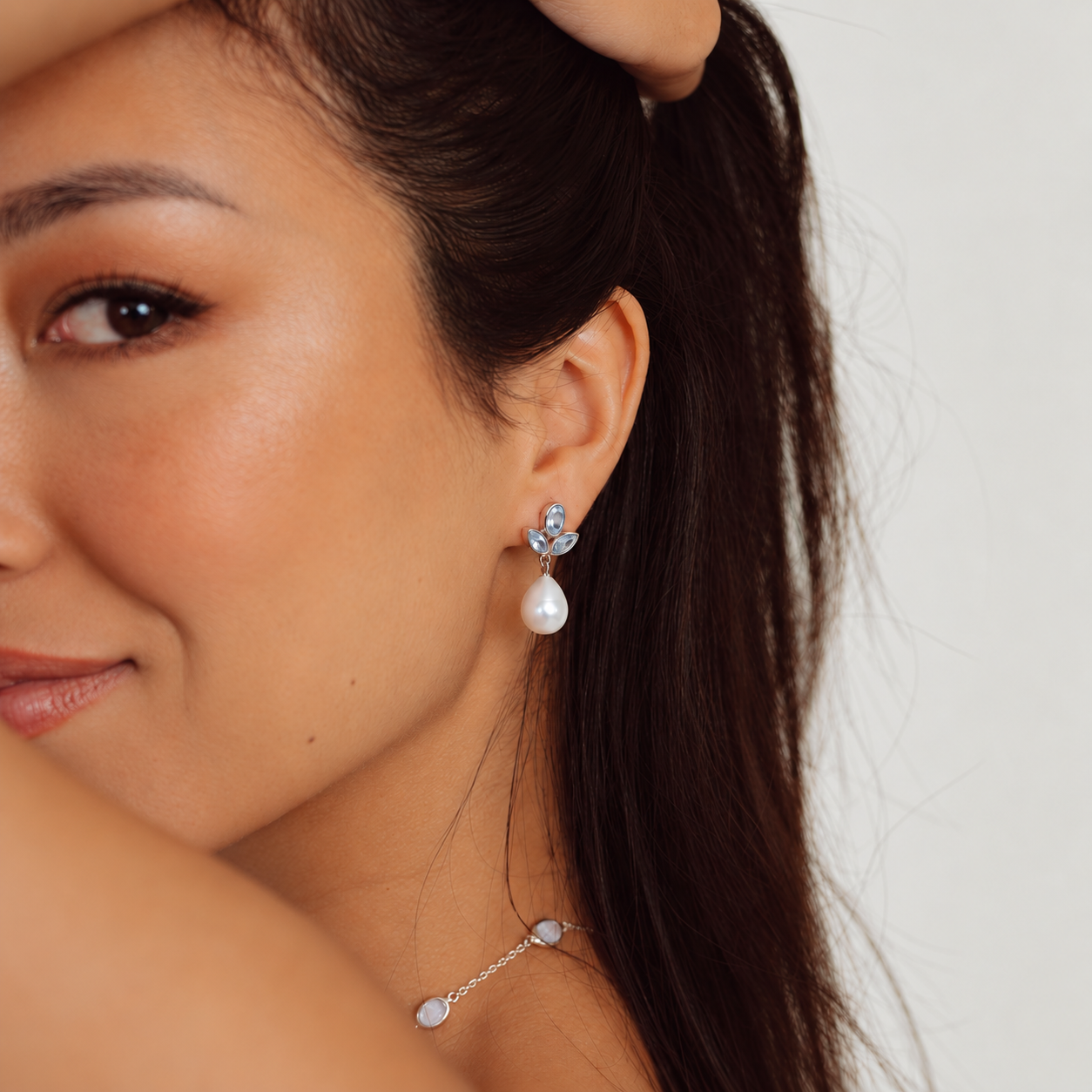Memara Jewellery. A close-up of a woman wearing a silver-toned dangle earring with a white teardrop pearl and a small clear gemstone, paired with a matching silver chain necklace featuring pale round beads.